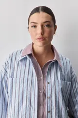 Camisa de calce relajado con diseño a rayas verticales en tonos azul y blanco, cuello clásico, mangas largas con puños abotonados y cierre frontal con botones. Presenta bolsillos aplicados en el delantero y una silueta versátil.