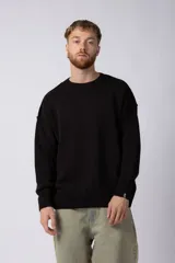 Sweater de punto negro con diseño oversized, cuello redondo y mangas largas con caída relajada. Presenta un tejido grueso y puños acanalados con una pequeña etiqueta distintiva en la manga.