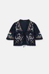 Top corto de color negro con escote en pico y mangas cortas anchas. Presenta bordados coloridos en estilo geométrico y floral, detalle de entredoses y cierre frontal mediante lazos con abalorios.