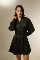 Vestido corto de satén negro, con cuello mao y detalle de volados en la botonadura frontal. Posee mangas largas con puños ajustados y un cordón fino en la cintura para entallar, terminado con pequeñas cuentas doradas.