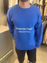 Sweater azul de hilo con cuello redondo, manga larga y puños y dobladillo acanalados. Presenta un estampado frontal con el texto "Bohemian Dept." y "Only Good News" en blanco.