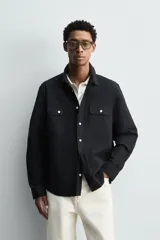 Sobrecamisa de corte relajado confeccionada en tejido ligero de algodón color negro. Presenta cuello de solapa, manga larga con puños abotonados y dos bolsillos de plastrón con solapa en el pecho. Cuenta con cierre frontal de botones a contraste y aberturas laterales en el bajo.