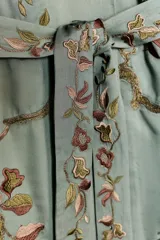 Kimono de largo midi en color verde grisáceo, con cuello de solapas amplias en pelo sintético marrón. Presenta mangas largas acampanadas y un cinturón ajustable del mismo tejido. El diseño está decorado con un delicado bordado floral en tonos tierra y rosados.