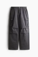 Pantalon cargo gris oscuro de corte holgado y diseño amplio, confeccionado en tejido de algodón. Cuenta con cintura elastizada con botón a presión, bolsillos laterales al bies y bolsillos cargo con solapa y botones a presión en las piernas.