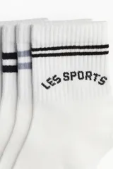 Pack de cinco pares de medias blancas de algodón con puño acanalado y borde superior elástico. Incluye diseños con rayas horizontales en negro y gris, y con la inscripción 'LES SPORTS' en negro.