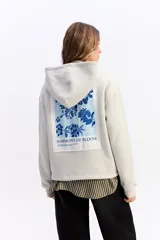 Canguro de algodón color beige con capucha amplia y corte relajado. Presenta un estampado rectangular en la espalda con motivos florales en tonos azules y el texto 'Harmony of Bloom'.