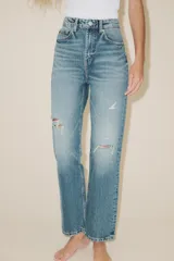 Jeans de tiro alto con diseño mom fit, confeccionados en denim de lavado medio con efecto desgastado y roturas localizadas. Presentan cinco bolsillos, trabillas para cinturón y cierre frontal con cremallera y botón metálico.