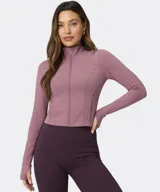 Campera Fabletics modelo Perfect Storm Cropped, de color rosado con cierre frontal completo, cuello alto, mangas largas con aberturas para pulgares y bolsillos laterales.