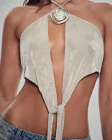 Top satinado color beige con diseño halter, escote profundo y detalle de dije metálico circular en el cuello. Presenta un nudo frontal y corte irregular.