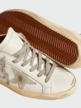 Zapatillas Golden Goose modelo Super Star, color blanco con estrella de gamuza gris, talón de cuero marrón y suela gastada.
