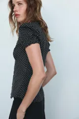 Camisa negra con estampado de lunares blancos, cuello redondo con escote en pico, mangas cortas abullonadas y detalle de volantes frontales. Cierre frontal con botones.