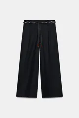 Pantalón de popelina de tiro alto y pernera ancha, de color negro. Presenta pinzas delanteras, bolsillos laterales y falsos bolsillos de vivo en la espalda. Incluye un cinturón combinado tipo pañuelo trenzado con tiras colgantes y cuentas de madera. Cierre frontal con cremallera, botón interior y gancho.