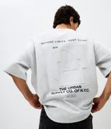 Remera gris melange de corte relaxed, con cuello redondo y mangas cortas. Presenta un diseño gráfico en la espalda con estampado y textura en alto relieve embossed que incluye la frase "Beyond Limits: Keep Going" y el texto "The Urban Supply Co. of N.Y.C.".