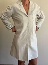 Camisa tipo vestido de corte recto y largo sobre la rodilla, confeccionada en viyela de algodón a rayas verticales. Presenta cuello solapa, cierre frontal con botones y mangas largas. Destaca por los detalles en tela estampada floral en los puños y en el bolsillo superior.