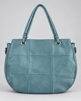 Cartera color celeste de diseño amplio y base redondeada, con textura granulada, costuras geométricas, doble asa de hombro y detalles metálicos con cierres.