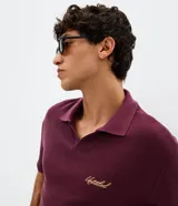 Remera de corte boxy color bordó, confeccionada en algodón con textura. Presenta cuello polo abierto sin botones, mangas cortas y bordado con lettering en el pecho.