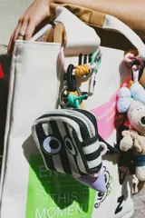 Bolsita cuadrada de tela con estampado de rayas verticales verdes y blancas, decorada con un aplique bordado de ojos grandes. Cuenta con cierre superior y una argolla metálica para colgar.