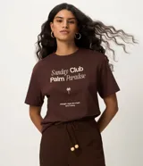 Remera marrón de manga corta y cuello redondo, confeccionada en algodón con estampa frontal que incluye tipografía "Sunday Club Palm Paradise", un icono de palmera y texto adicional.