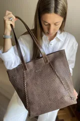 Bolso grande tipo tote color beige con textura tejida y costuras decorativas en los laterales. Tiene doble asa de hombro y logo de la marca en el frente. Interior forrado con bolsillo con cierre y portacelular.