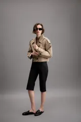 Trench corto de color beige con cuello de solapa, cierre frontal cruzado con botones y mangas largas con trabillas ajustables en los puños. Presenta un diseño estructurado con hombreras y bolsillos delanteros de vivo.