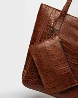 Bolso shopper negro de cuero con textura de cocodrilo, doble asa de hombro y dije colgante rectangular.