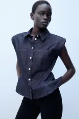 Camisa sin mangas de denim azul oscuro, con cuello solapa, hombreras marcadas, bolsillos delanteros de plastrón, bajo con peplum y cierre frontal con botones metálicos.