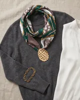Pañuelo de seda estampado con diseño de cadenas, lunares y detalles geométricos en tonos verde, marrón, blanco y dorado. Incluye un dije ovalado dorado con textura martillada.
