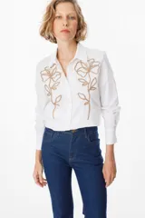 Camisa blanca de manga larga con cuello clásico y cierre frontal de botones. Presenta un delicado bordado lineal de flores en color visón sobre ambos lados del frente.