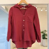 Camisa de lino color bordó, de manga larga, con cuello camisero, cierre frontal con botones nacarados, bolsillo aplicado en el pecho y diseño de corte asimétrico con la parte posterior más larga.