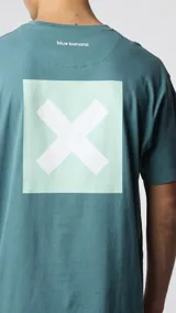 Camiseta de manga corta color verde azulado, corte holgado, con un pequeño estampado cuadrado con una 'X' en el pecho.