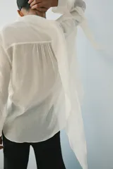 Camisa de hilatura con mezcla de lyocell color blanco, con cuello fluido con pañuelo incorporado, manga larga con puño y bajo redondeado asimétrico. Cierre frontal con botones ocultos.