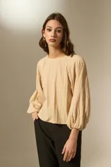 Blusa de color beige con cuello redondo y mangas largas abullonadas con puños elásticos. Presenta un corte holgado y fluido.
