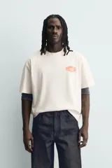 Camiseta de corte relajado color crudo, con cuello redondo y manga corta. Presenta un estampado gráfico pequeño en el pecho y un diseño grande en la espalda relacionado con el artista Post Malone.