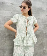Blusa estampada con hojas verdes y blancas, con escote en V y mangas cortas abullonadas. Presenta volados en el ruedo y detalle de lazos en el frente.