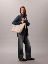 Cartera tote Calvin Klein de color beige, confeccionada en suave piel sintética, con diseño estructurado, asas fijas y compartimentos exteriores en los laterales. Presenta detalles de cinta con logo en jacquard y logo metálico grabado en la parte delantera.