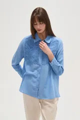 Camisa de manga larga color celeste, con cuello camisero y cierre frontal de botones. Presenta un diseño distintivo con paneles de encaje tipo crochet en la parte superior delantera.