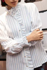 Camisa de manga larga con estampado de rayas verticales celestes sobre fondo blanco. Presenta un cuello alto con volados, cierre frontal con botones y puños con volados.