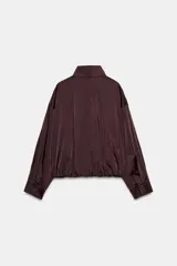 Cazadora satinada de color negro, con cuello subido, manga larga y bolsillos frontales de plastrón con cordones decorativos. Cuenta con bajo elástico y cierre frontal con cremallera y botones a presión oculta.