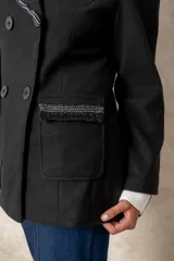 Blazer cruzado color beige, con cierre de cuatro botones frontales. Presenta detalles artesanales de encaje con perlas y cristales en la solapa y en los bolsillos delanteros.