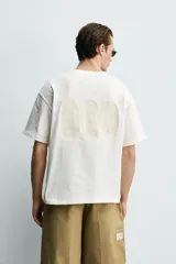 Camiseta de corte relajado, color crudo, con cuello redondo y manga corta. Presenta un diseño con texto estampado y bordados a contraste en la parte frontal.
