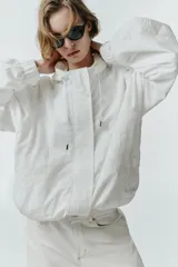 Chaqueta blanca de cuello redondo con capucha ajustable mediante cordones elásticos. Presenta manga larga con puños elásticos, bolsillos delanteros y bajo asimétrico ajustable con cordones y stoppers. El cierre frontal combina cremallera y botones ocultos bajo una solapa.