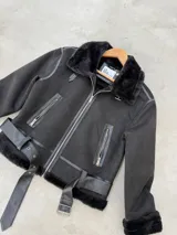Campera tipo gamulán de corte aviador, color negro, confeccionada en gamuza sintética con interior de piel sintética. Presenta cierre frontal, bolsillos laterales con cierre, detalles en cuero sintético y cinturón ajustable en la cintura.