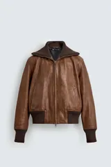 Cazadora de cuero color marrón, estilo bomber, con cuello ancho de punto acanalado marrón oscuro, cierre frontal de cremallera y puños y cintura elásticos de punto acanalado marrón oscuro.