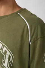 Remera de corte oversize color verde oliva con estampado frontal estilo universitario y detalles de líneas blancas en los hombros y laterales.
