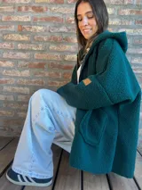 Campera de corderito color verde oscuro, con diseño holgado, cuello alto y manga larga. Presenta una etiqueta de cuero sintético en la manga.