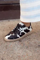 Zapatillas bajas con estampado de vaca en blanco y negro, cordones negros y suela color caramelo.