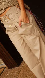Pantalón ancho de tiro alto color beige, con pliegues frontales y botones visibles en la cintura. El ruedo del pantalón tiene un detalle de terminación en cuero color caramelo.