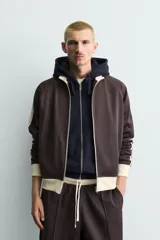 Sudadera de corte relajado en color azul marino con cuello alto y cierre frontal de cremallera. Presenta bandas laterales en color crema a contraste que recorren las mangas, puños y bajo en tejido rib, y bolsillos laterales de vivo.