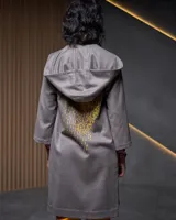 Túnica gris con capucha, confeccionada en gabardina de lana estampada artesanalmente con diseño abstracto en tonos ocre. Presenta dos piezas a modo de bufandas que se anudan en la parte delantera, bolsillos laterales y capucha amplia.