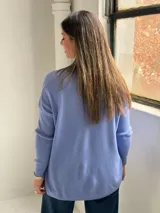 Sweater de punto color azul lavanda, con escote en V, mangas largas con puños acanalados y dos bolsillos frontales tipo parche. Presenta una costura central vertical visible.